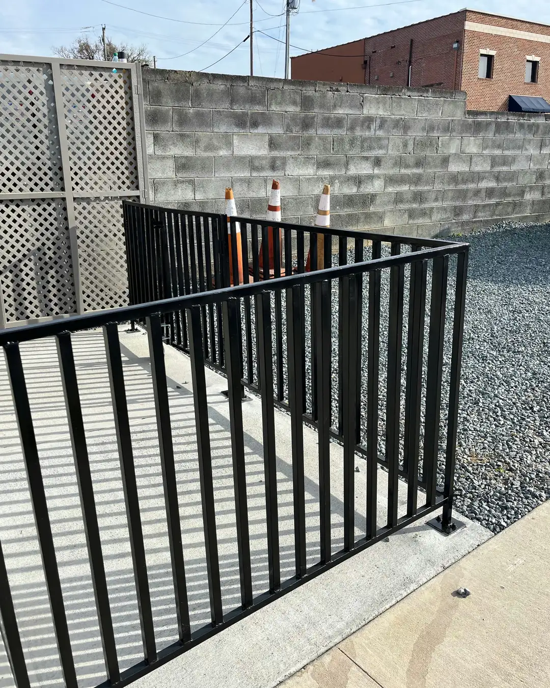A black metal railing separating a concrete path from a gravel area with a cinder block wall and brick buildings.
